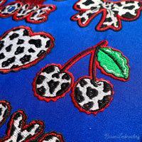 Two cherries with cow print texture and green leaves, finished with a wavy red border embroidery design.