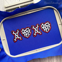 Machine embroidery design of the letters XOXO where both O characters are replaced by cow print hearts with wavy red borders.