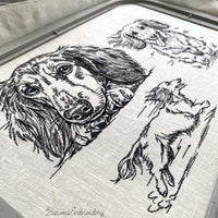 Black and white embroidered illustration of a long-haired dachshund dog. The dog is lying down, with its paws and head resting on a surface. The dog's eyes are open, and it has long, flowing fur.