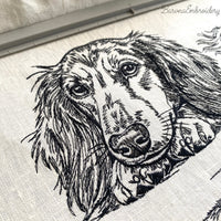 Black and white embroidered illustration of a long-haired dachshund dog. The dog is lying down, with its paws and head resting on a surface. The dog's eyes are open, and it has long, flowing fur.