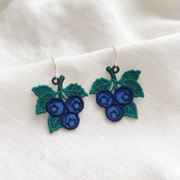 Pair of blue Blueberry FSL earrings with detailed green leaves and silver hooks displayed on soft white fabric