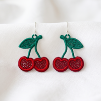Pair of red cherry FSL earrings with green leaves and silver hooks displayed on soft white fabric