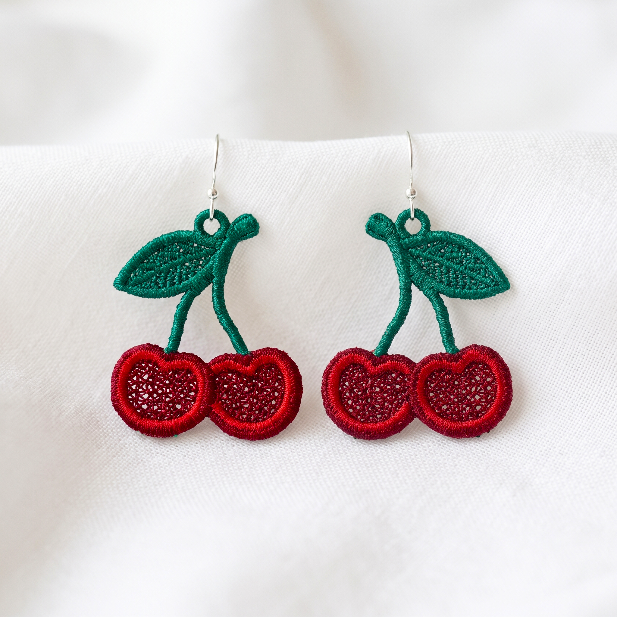 Pair of red cherry FSL earrings with green leaves and silver hooks displayed on soft white fabric