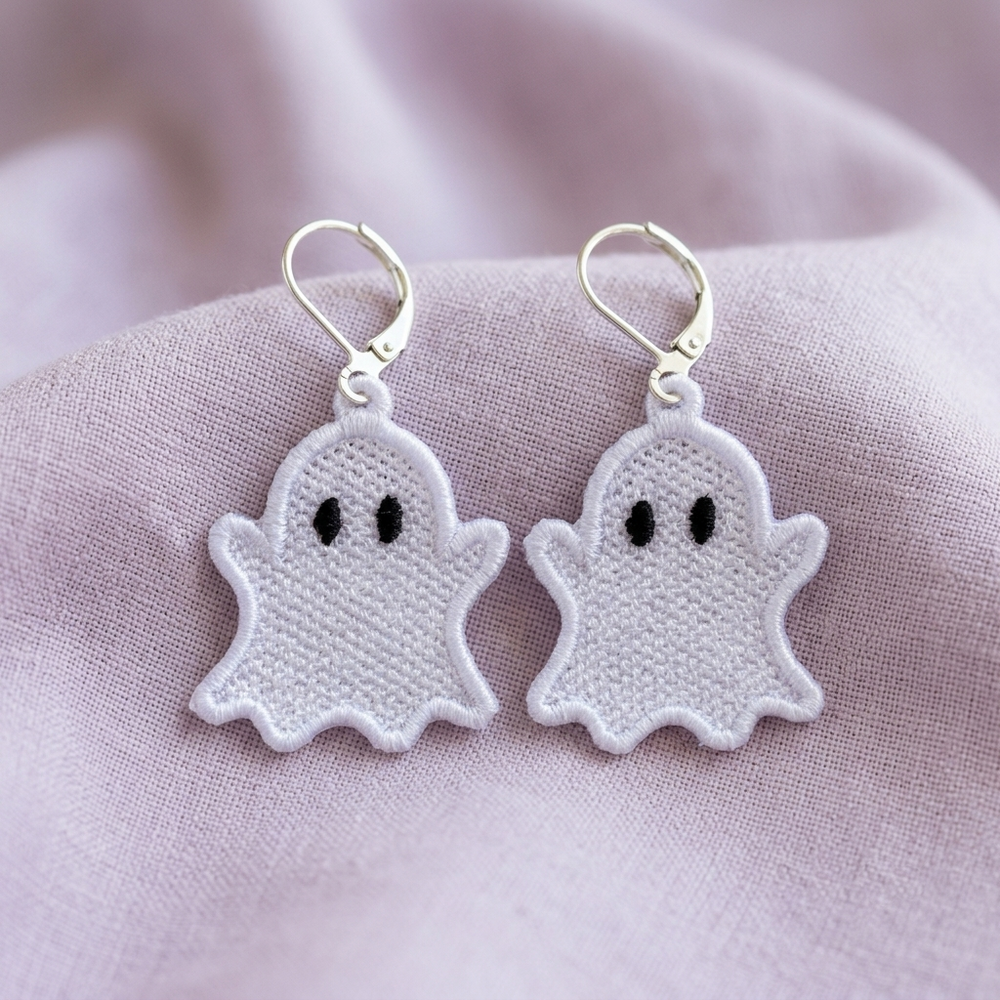 Pair of white ghost FSL earrings with black oval eyes and silver hooks displayed on lavender fabric
