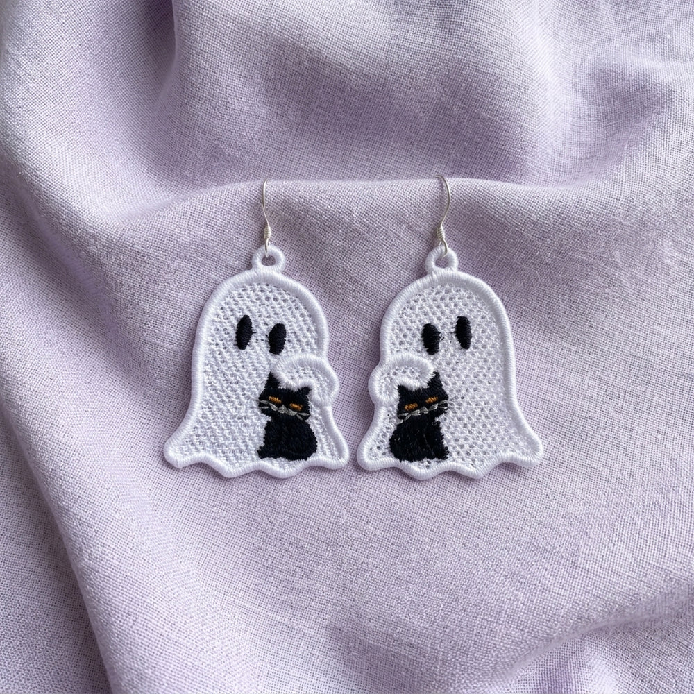 Pair of mirrored white ghost FSL earrings cuddling black cats with yellow eyes on soft lavender fabric