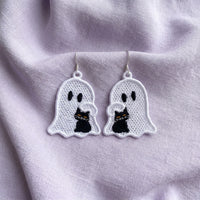 Pair of mirrored white ghost FSL earrings cuddling black cats with yellow eyes on soft lavender fabric