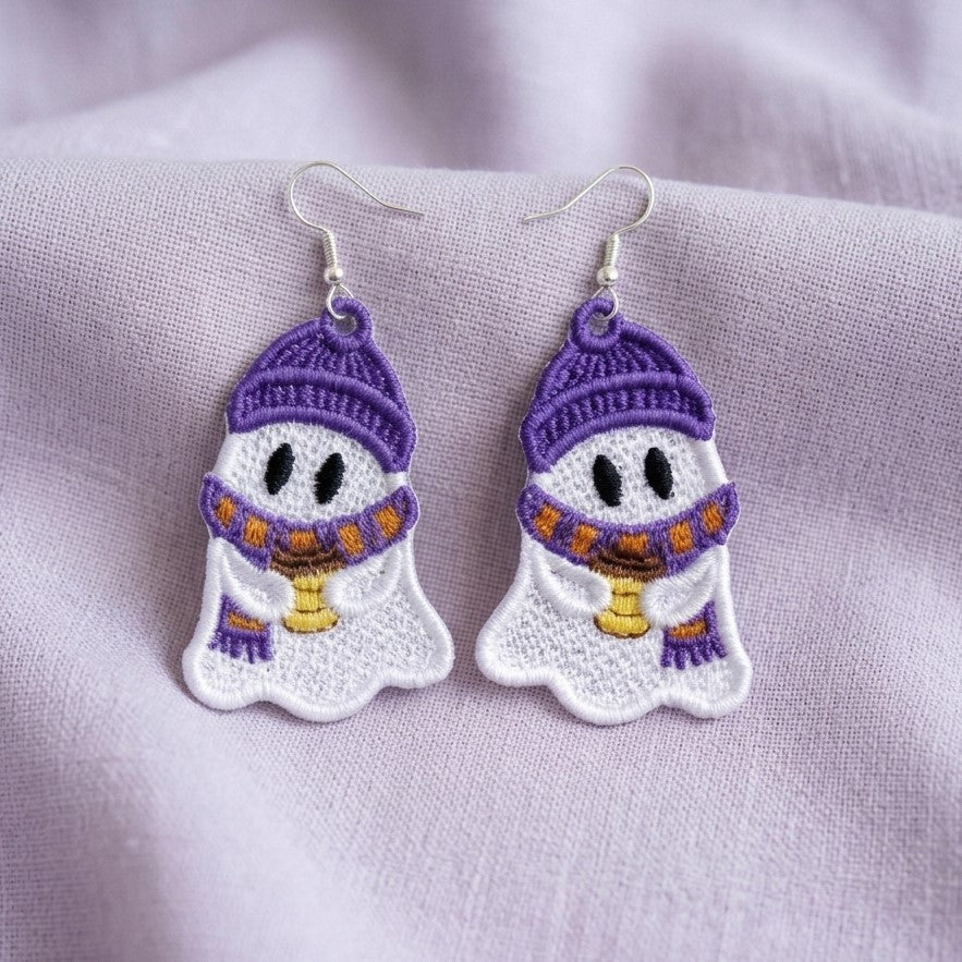 White ghost earrings with purple hats and scarves, holding coffee cups, displayed on soft lavender fabric.