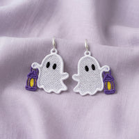 Pair of white ghost FSL earrings holding purple lanterns with yellow centers on soft lavender fabric