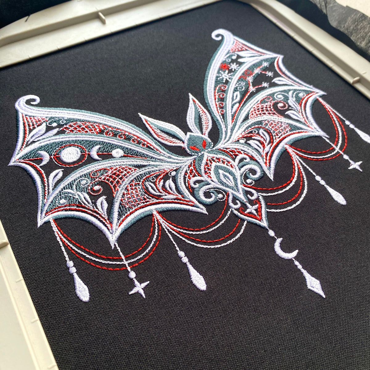 Gothic bat stitched on dark fabric in a hoop, with moon phases in one wing, stars in the other and pendant motifs below.
