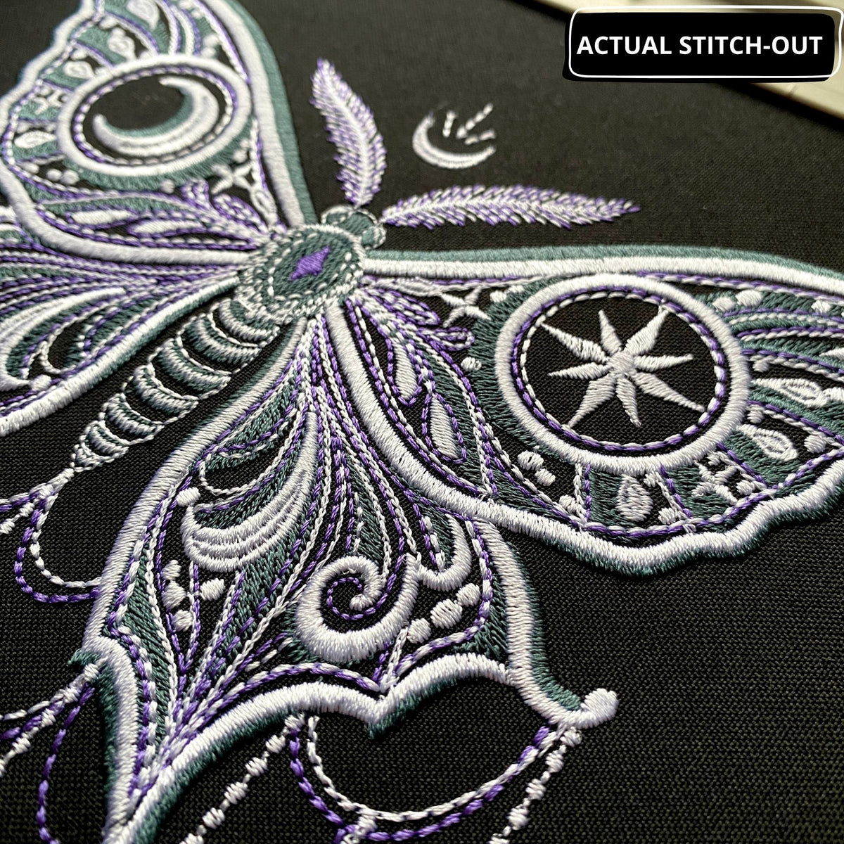 Actual gothic moon moth stitch-out on dark fabric in a hoop, with ornate wings and stitched chains ending in moons and stars.