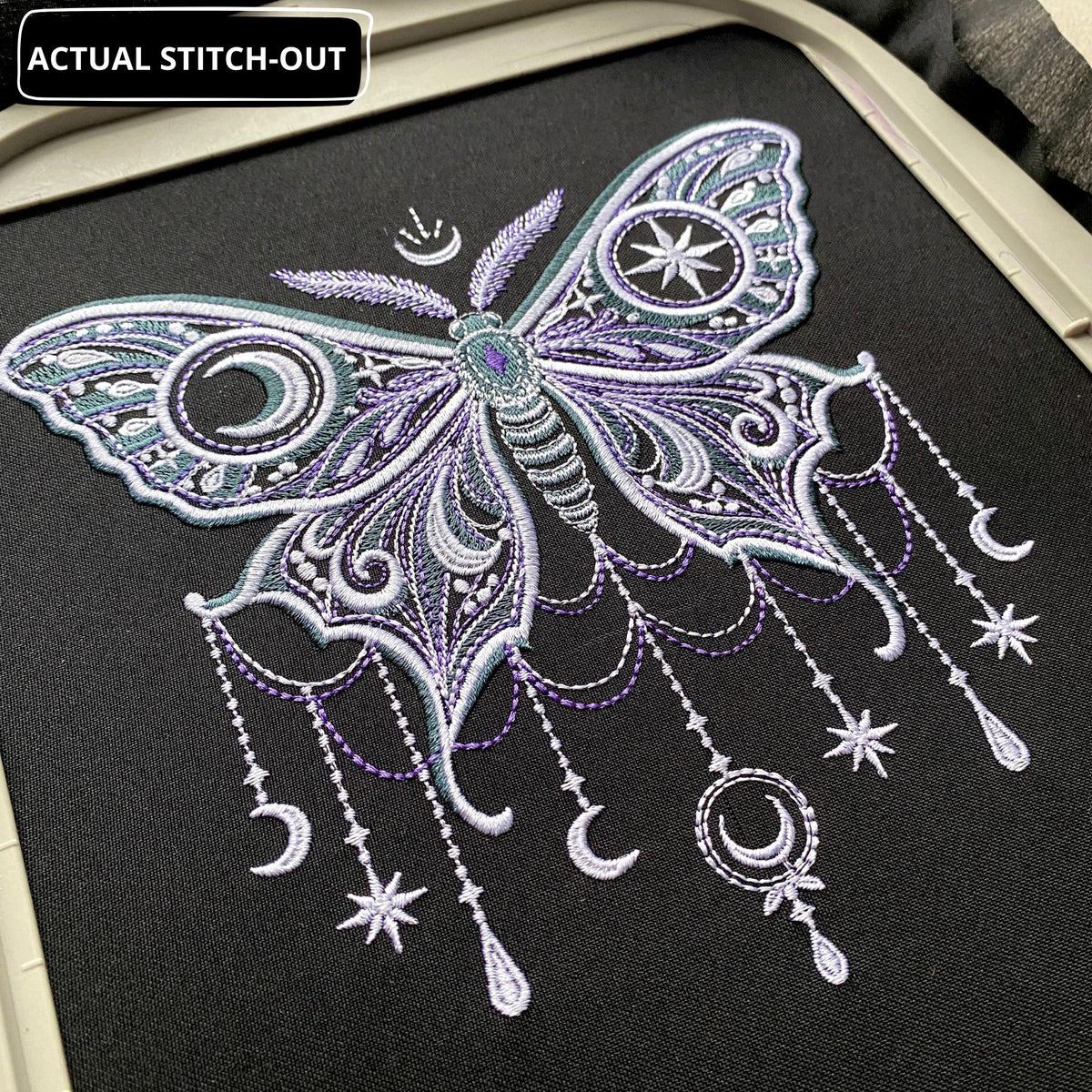Close-up of actual moon moth embroidery on dark fabric, showing pale satin outlines, purple stitching and a star within the wing.