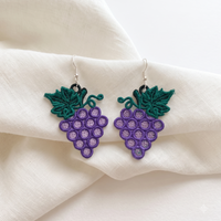 Pair of purple grape cluster FSL earrings with green vine leaves and silver hooks displayed on soft cream fabric