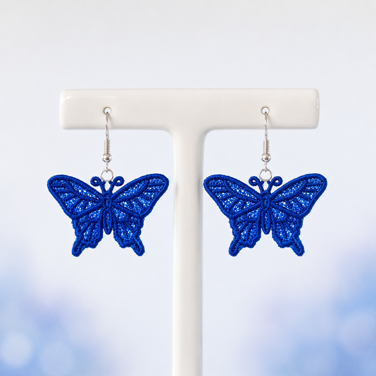 Pair of blue swallowtail butterfly FSL earrings with lace-filled wings and silver hooks on a white jewelry stand