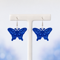 Pair of blue swallowtail butterfly FSL earrings with lace-filled wings and silver hooks on a white jewelry stand