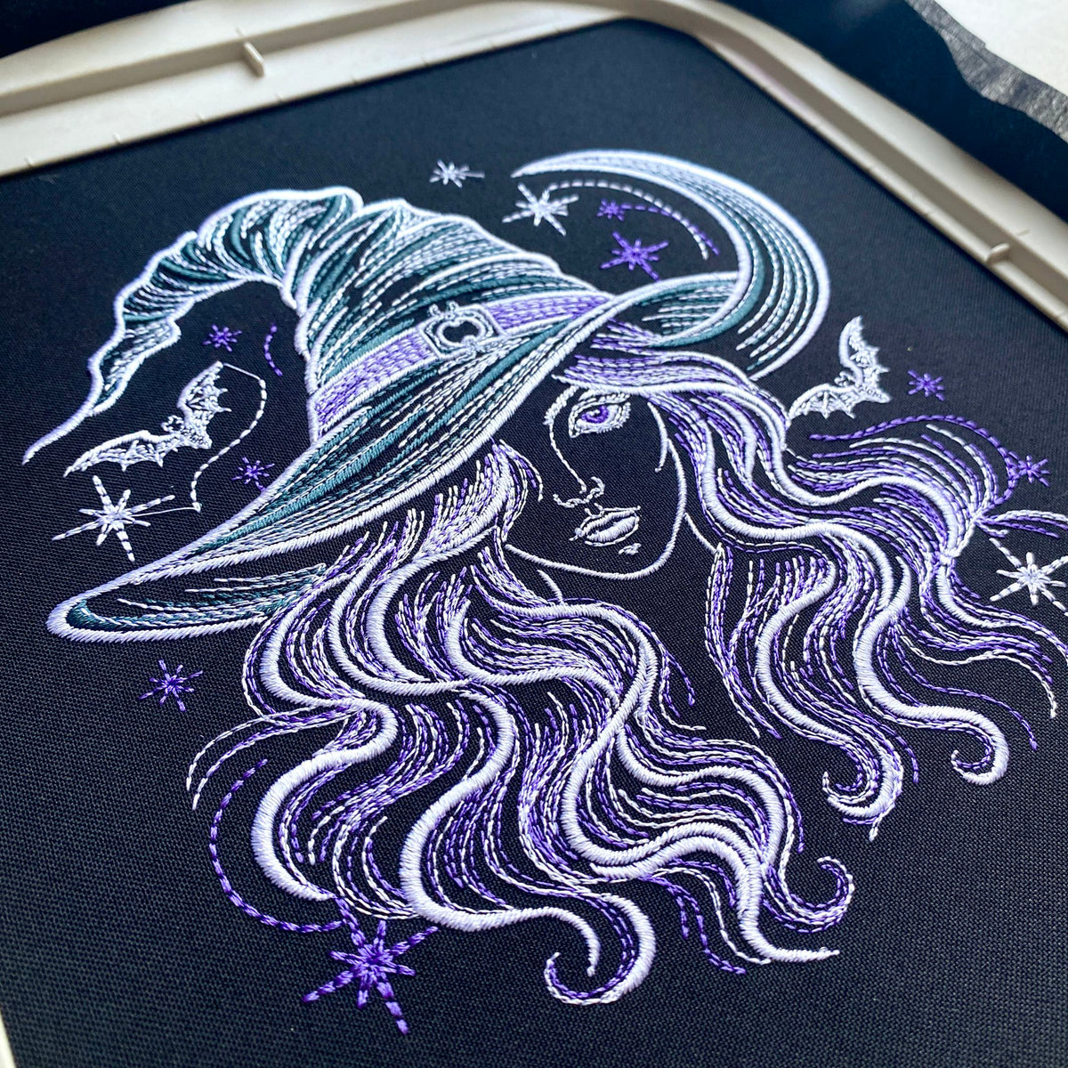 Witch portrait stitched on dark fabric in an embroidery hoop, surrounded by bats, stars and a crescent moon.