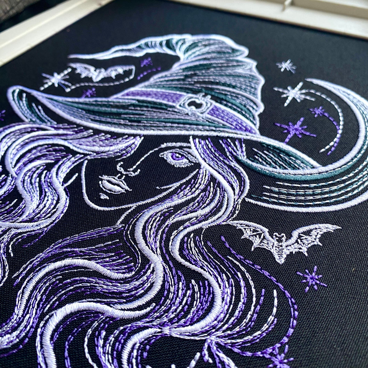 Close-up of witch embroidery on dark fabric showing satin outlines, purple hair details and crescent moon stitching.